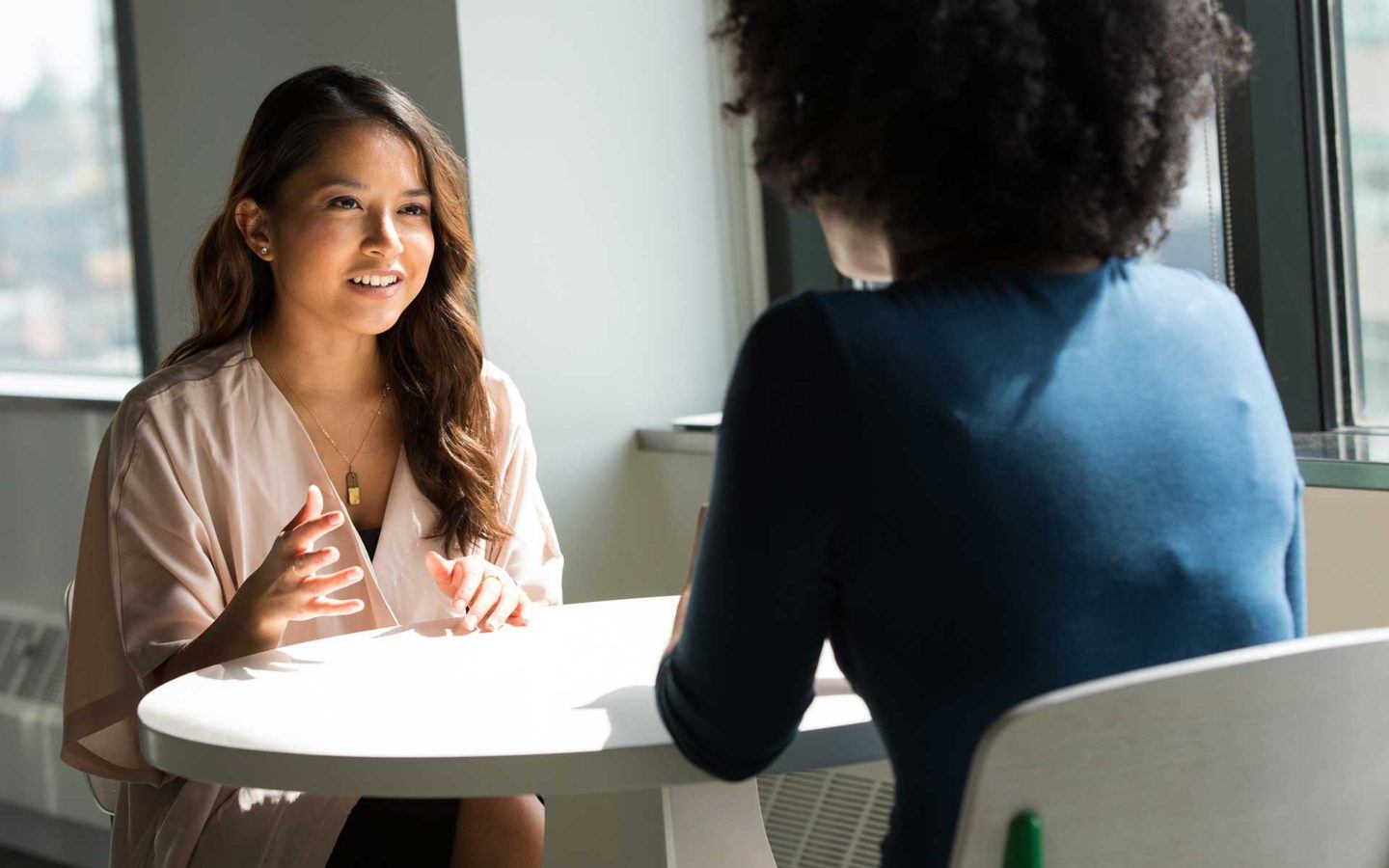 woman in beige dress is speaking with a woman in a blue shift at a table in an office