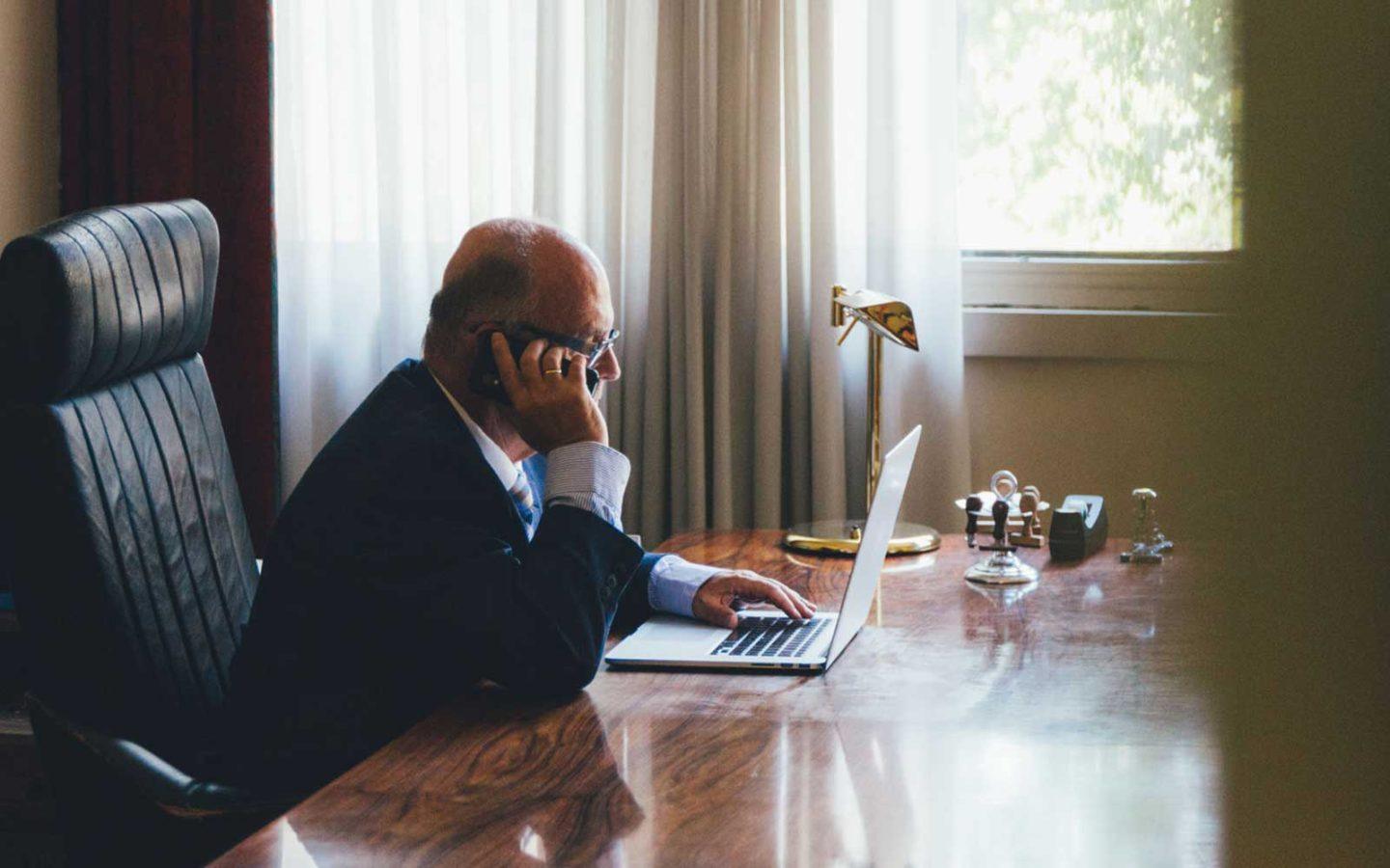 a man on a cell phone, sitting in front of a laptop working at a large wooden desk